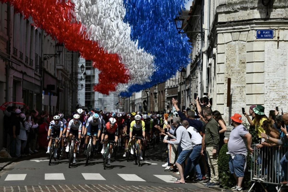 The pack of riders (peloton) cycles under decorative streamers hung above the race route in Aire-sur-la-Lys during the 3rd stage of the 112th edition of the Tour de France cycling race, 178.3 km between Valenciennes and Dunkerque (Dunkirk), Northern France, on July 7, 2025. Marco BERTORELLO / AFP